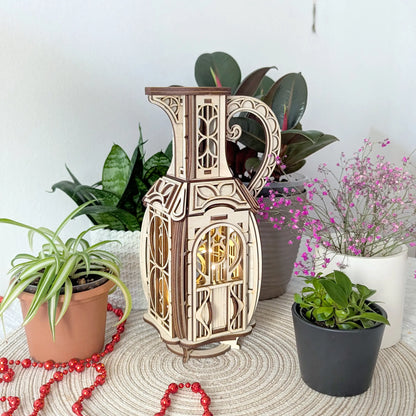 Assembled laser cut plywood jug fairy house glowing with warm internal light on a table surrounded by green houseplants.