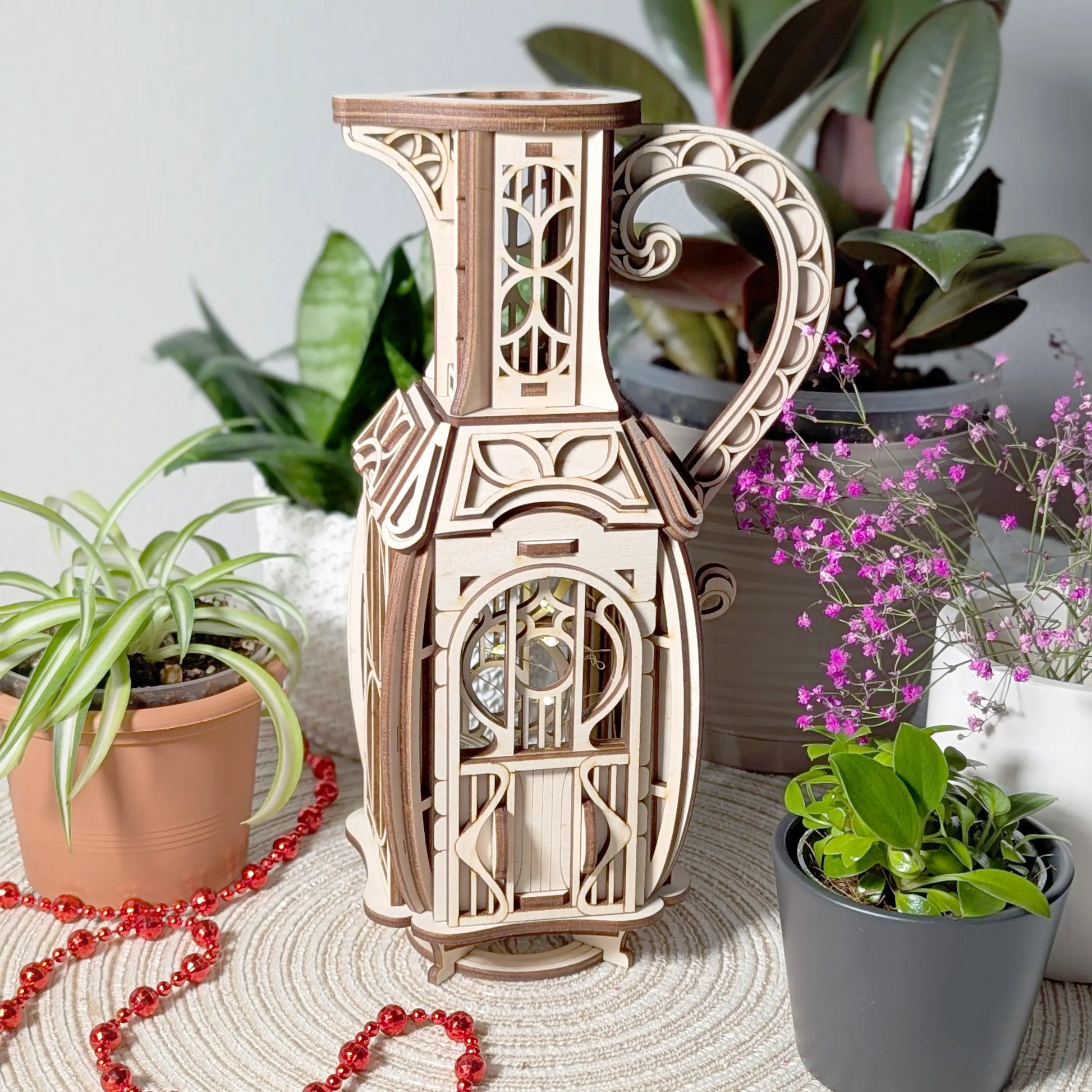 Full view of a whimsically shaped laser cut plywood jug fairy house styled on a table with indoor potted plants and beads.