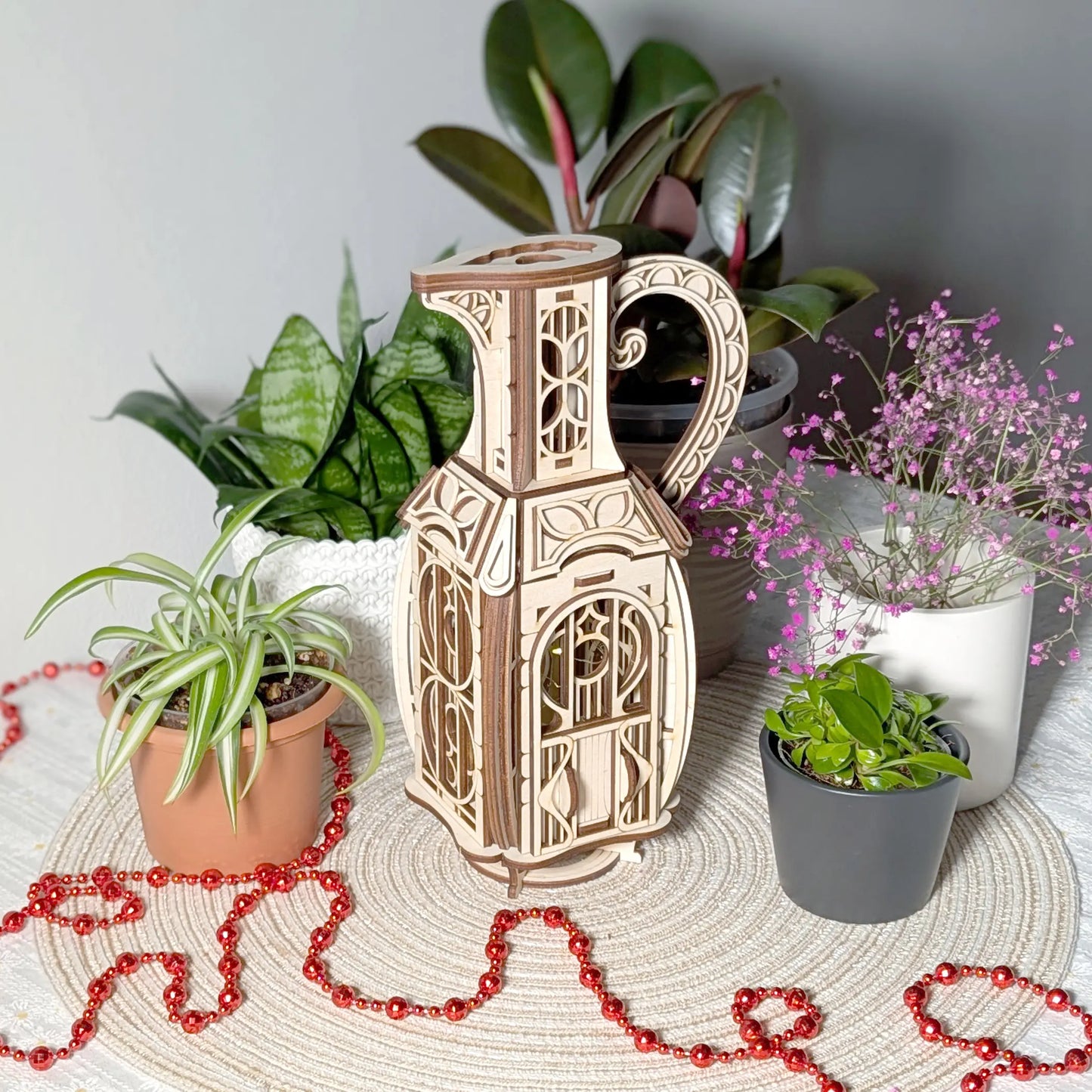 Finished laser cut plywood jug fairy house sitting on a woven mat among potted green plants and a red decorative garland.