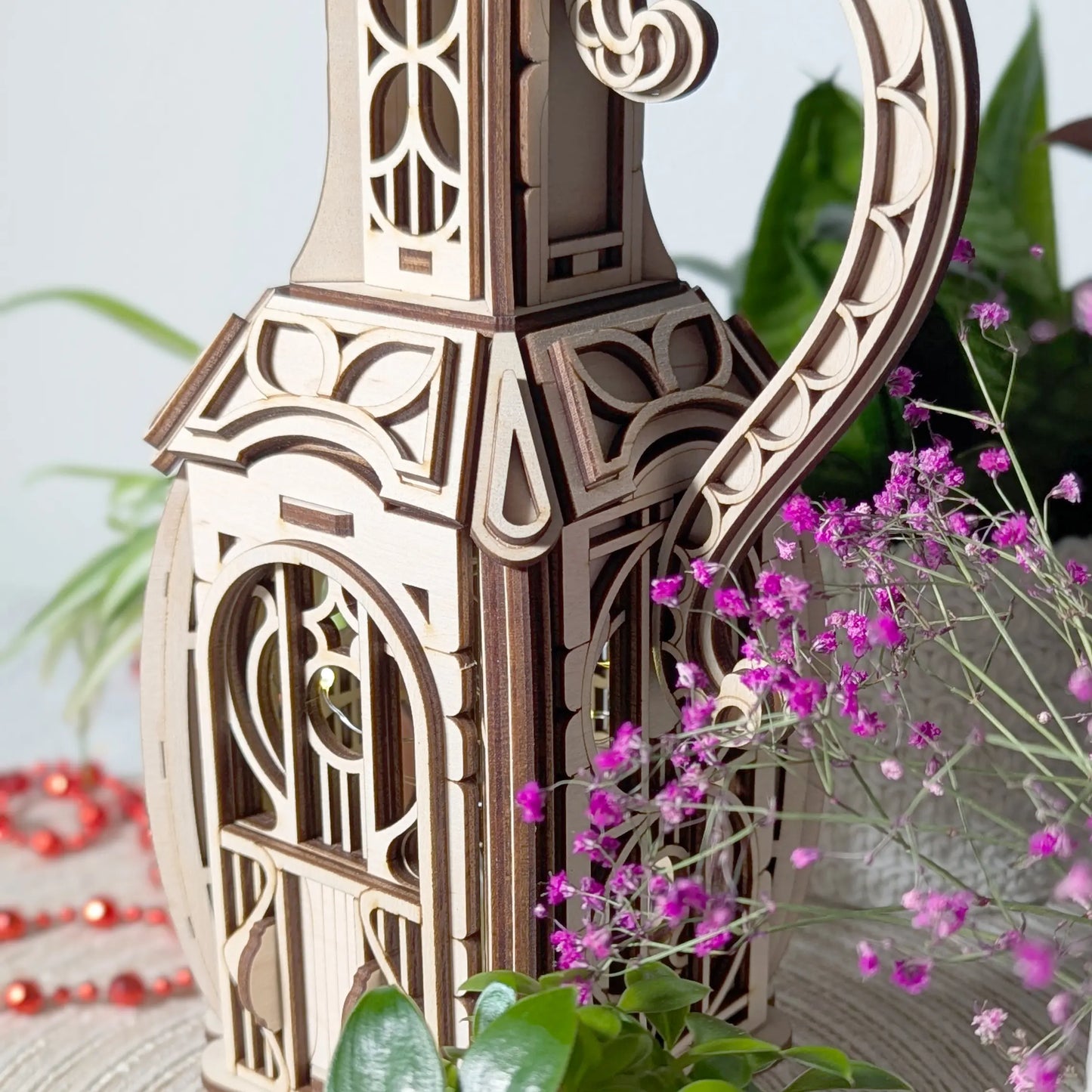 Macro close-up of a laser cut plywood jug house door partially framed by delicate pink flowers and green indoor plants.