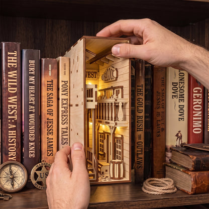 Hands inserting a lit laser-cut birch plywood Wild West saloon book nook into a bookshelf, showing natural wood grain.