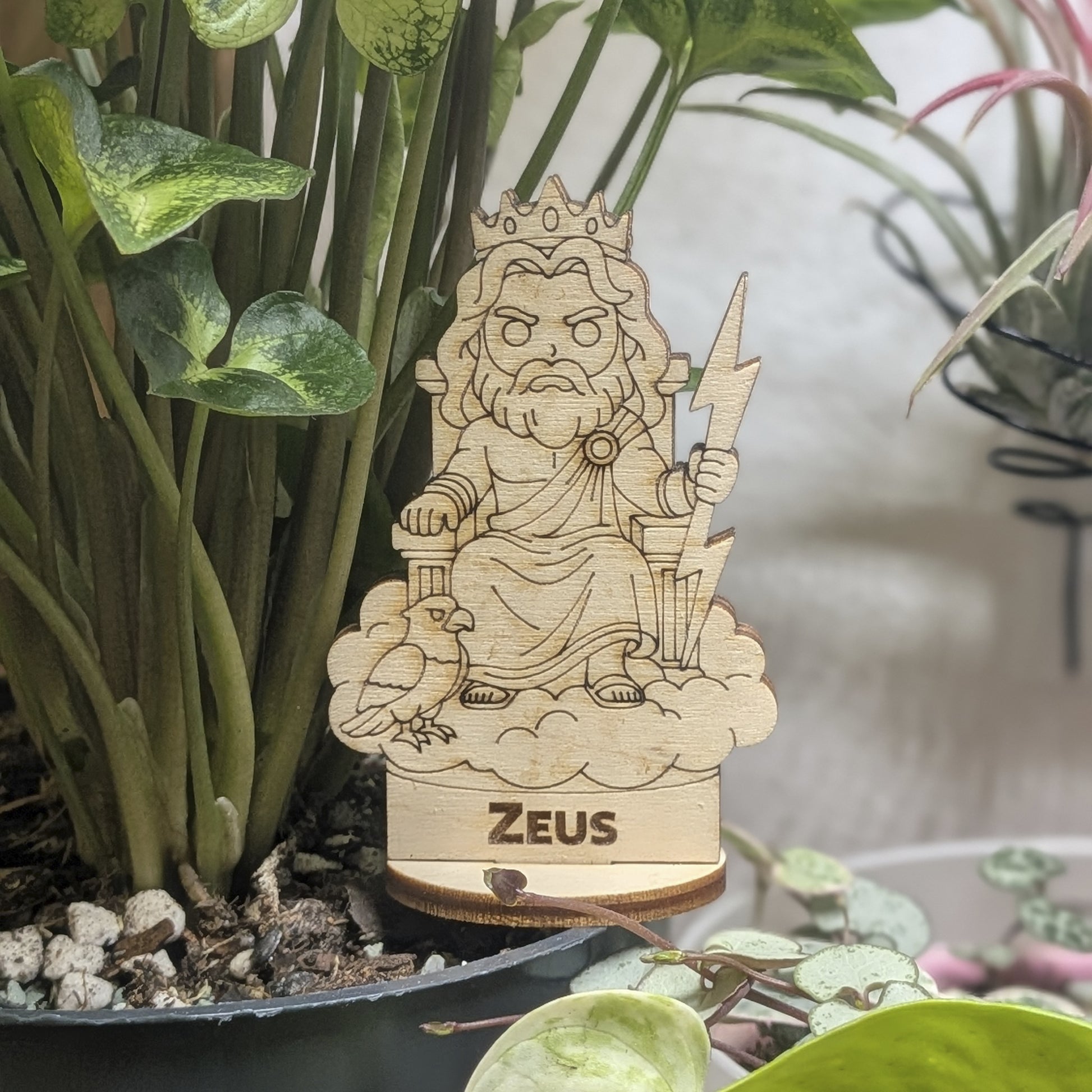 Laser cut plywood Zeus figurine sits in a plant pot. Features a lightning bolt, thick beard, and robes on a round wood base