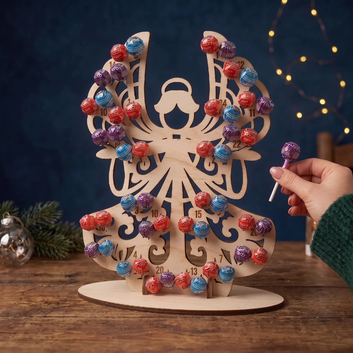 Hand removes lollipop from standing laser cut plywood angel advent calendar on wood table with fairy lights