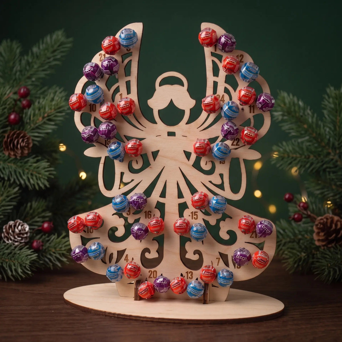 Laser cut plywood angel advent calendar holding 24 lollipops displayed on tabletop between evergreen branches.
