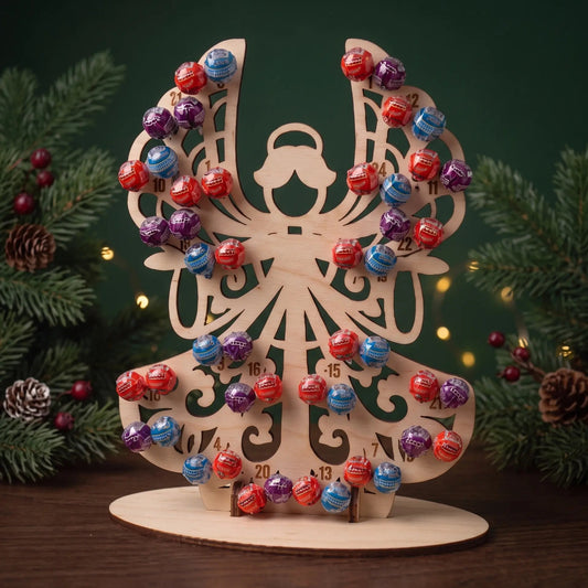 Laser cut plywood angel advent calendar holding 24 lollipops displayed on tabletop between evergreen branches.