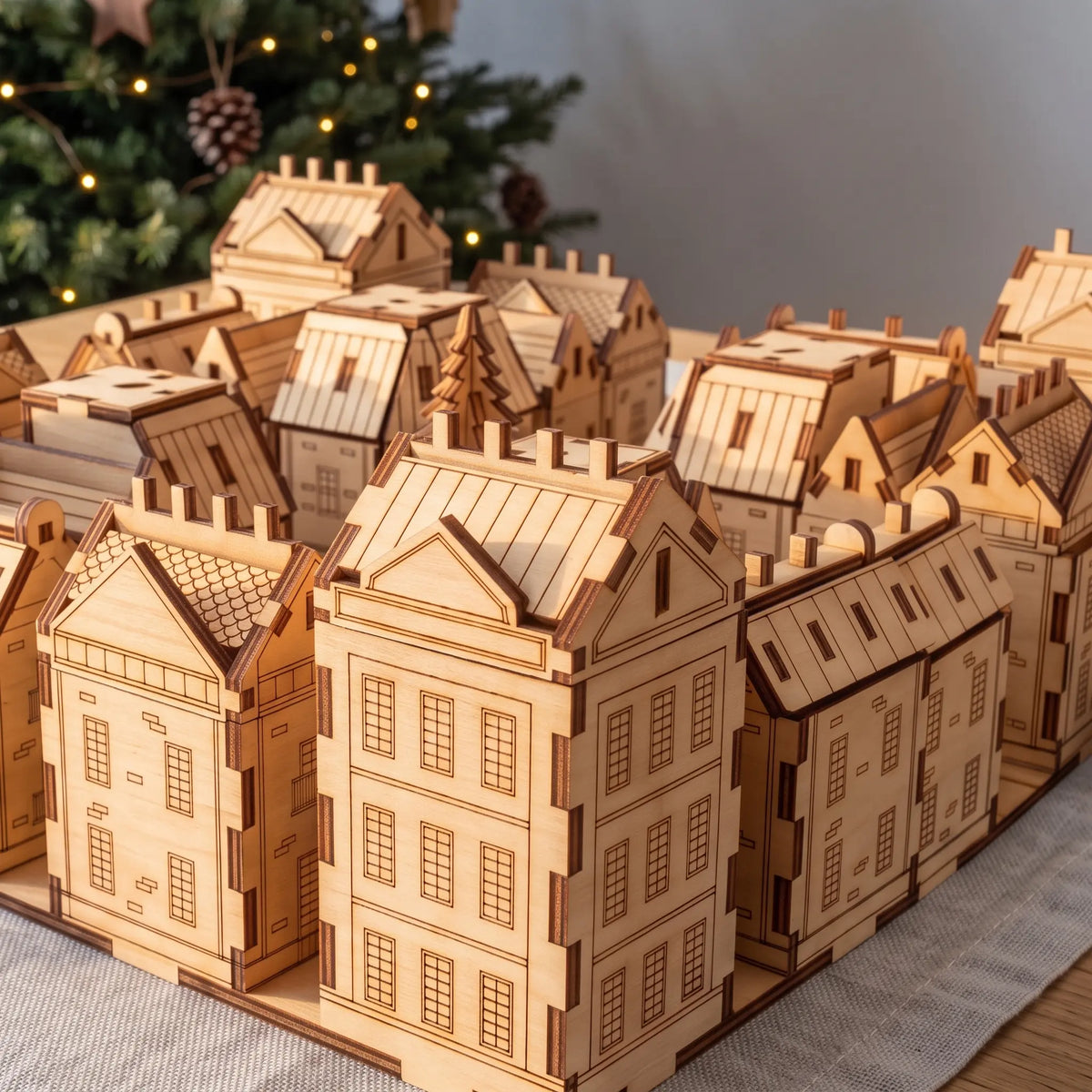 Laser cut plywood miniature town advent calendar standing on tabletop in front of decorated holiday Christmas tree.