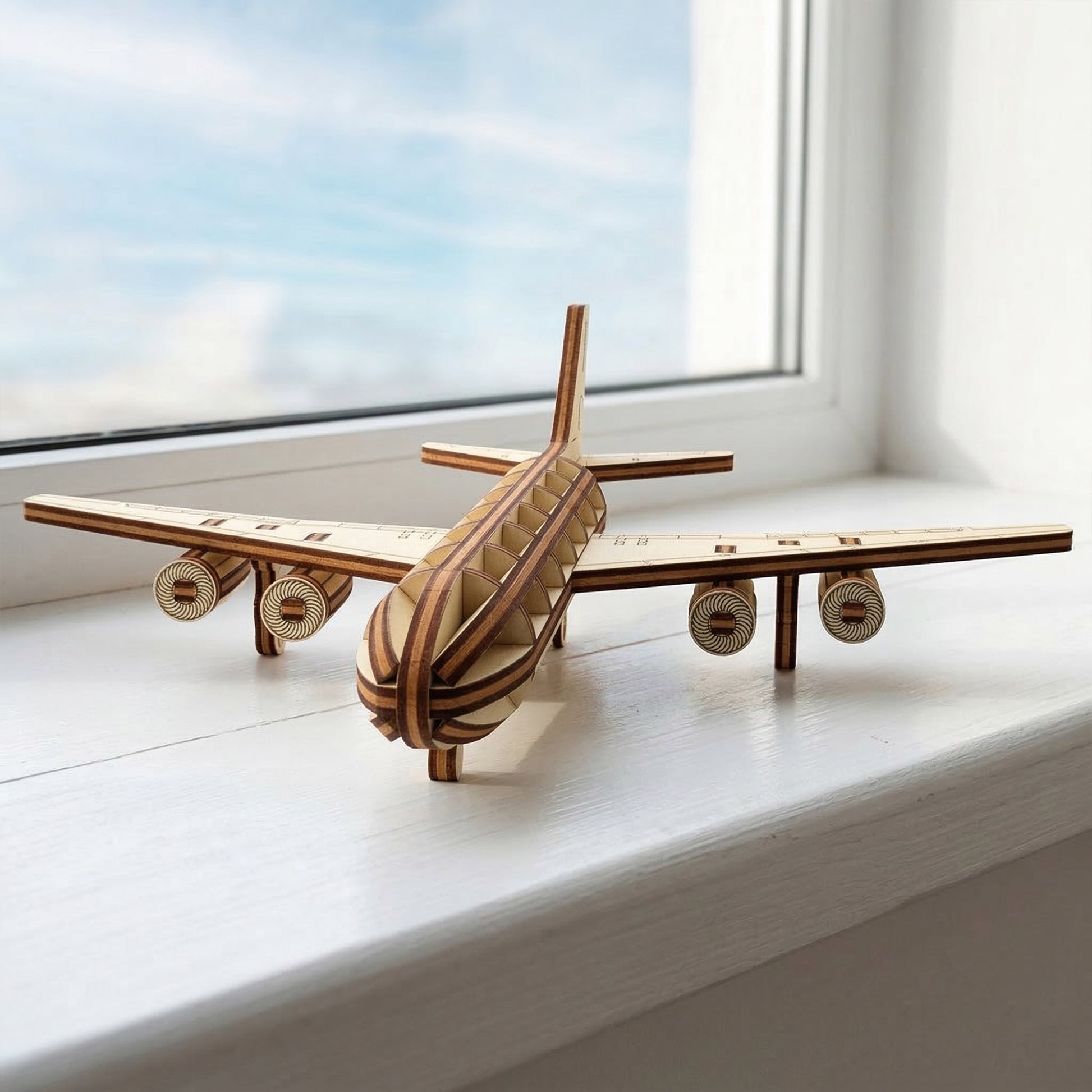 Front-facing view of a laser cut plywood passenger jet model. The symmetrical design highlights four detailed engines and a ribbed wooden body. The aircraft is resting on a white ledge with a bright blue sky visible through the window behind it.