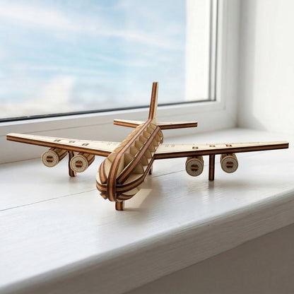 Front-facing view of a laser cut plywood passenger jet model. The symmetrical design highlights four detailed engines and a ribbed wooden body. The aircraft is resting on a white ledge with a bright blue sky visible through the window behind it.