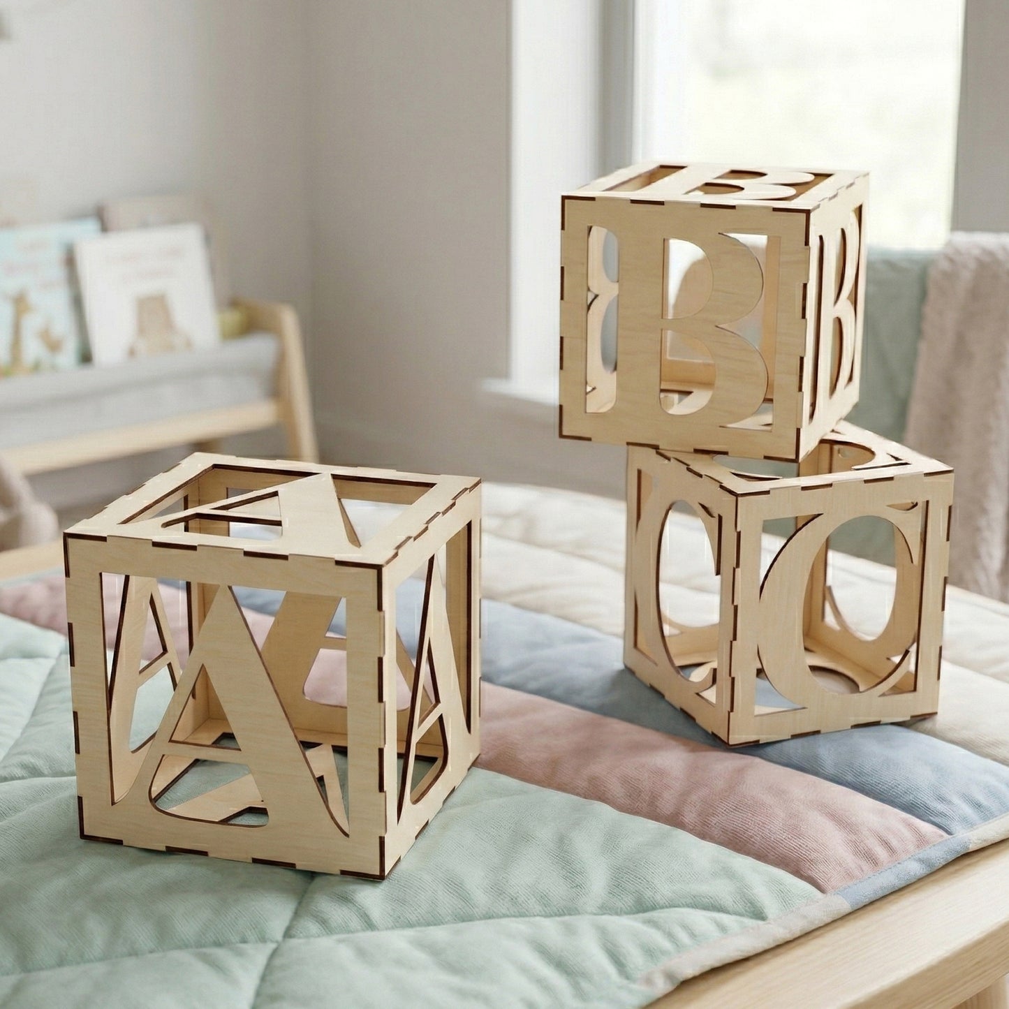 Three large hollow alphabet cubes (A, B, and C) made from laser-cut plywood. The blocks feature a finger-joint construction and bold, cut-out letters on all sides. Posed on a pastel quilted blanket in a bright, modern nursery setting.
