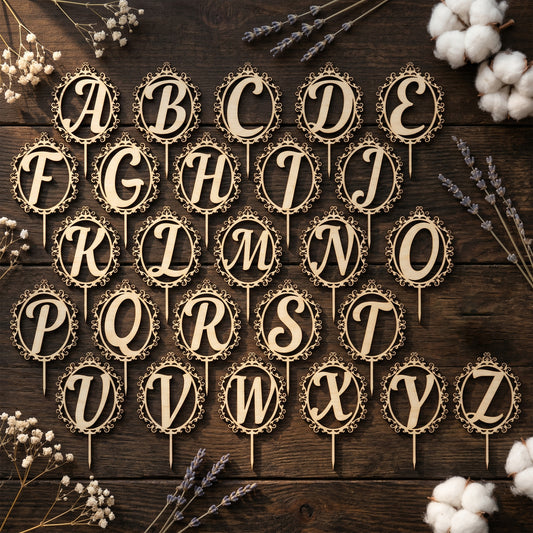 A complete set of 26 laser-cut plywood alphabet cake toppers featuring a vintage, ornate filigree design. Each wooden monogram topper shows a script letter from A to Z inside an intricate circular frame with a pointed stake for insertion. Displayed in a flat lay on a dark rustic wood surface with dried lavender, baby's breath, and cotton bolls.