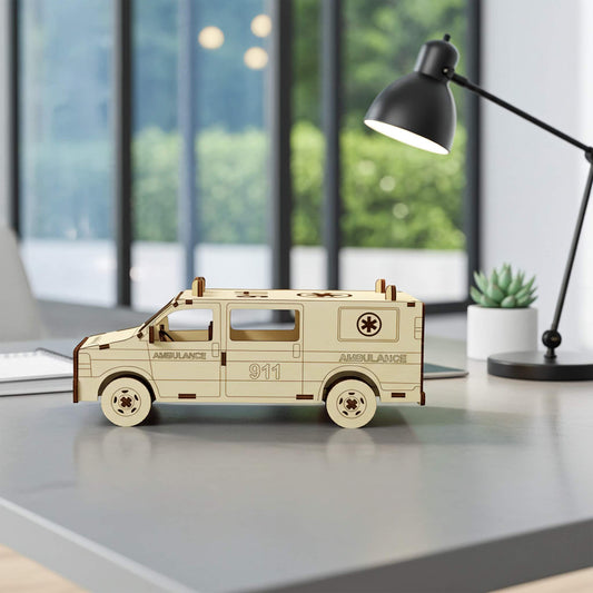 Side profile of a laser cut wooden plywood ambulance model sitting on a grey office desk. The 3D puzzle showcases precise window cutouts, wheels with star details, and engraved emergency markings. Background features a black desk lamp and a succulent