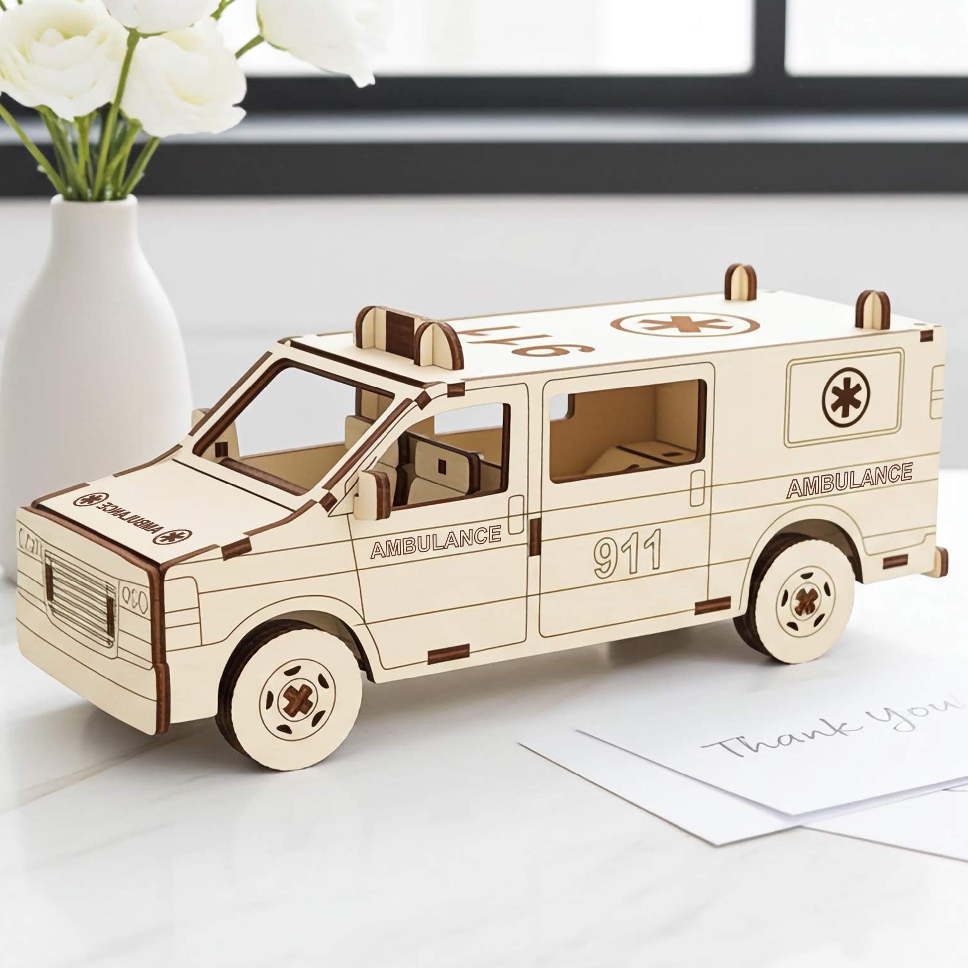 Laser cut wooden plywood ambulance 3D model on a white marble surface next to a vase of white ranunculus. Detailed plywood assembly featuring "911" and "AMBULANCE" engravings, roof lights, and a medical cross symbol. Clean, bright room background