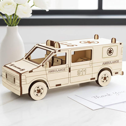 Laser cut wooden plywood ambulance 3D model on a white marble surface next to a vase of white ranunculus. Detailed plywood assembly featuring "911" and "AMBULANCE" engravings, roof lights, and a medical cross symbol. Clean, bright room background