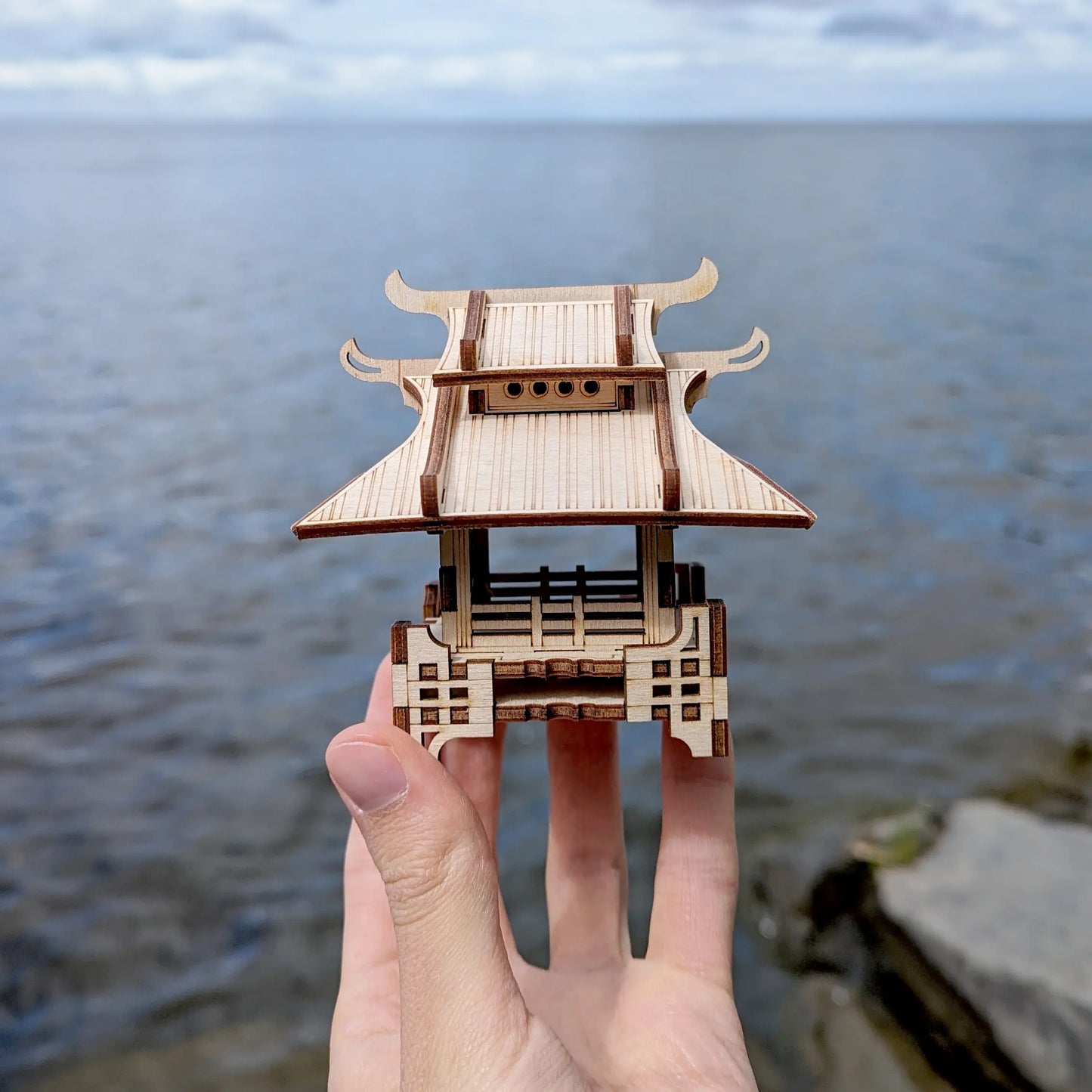 Close-up of a tiny laser cut plywood japanese pavilion held in a hand against a blurred blue sea horizon background.