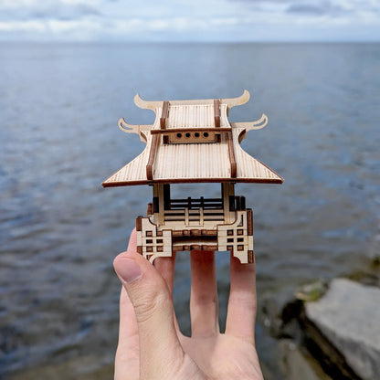 Close-up of a tiny laser cut plywood japanese pavilion held in a hand against a blurred blue sea horizon background.