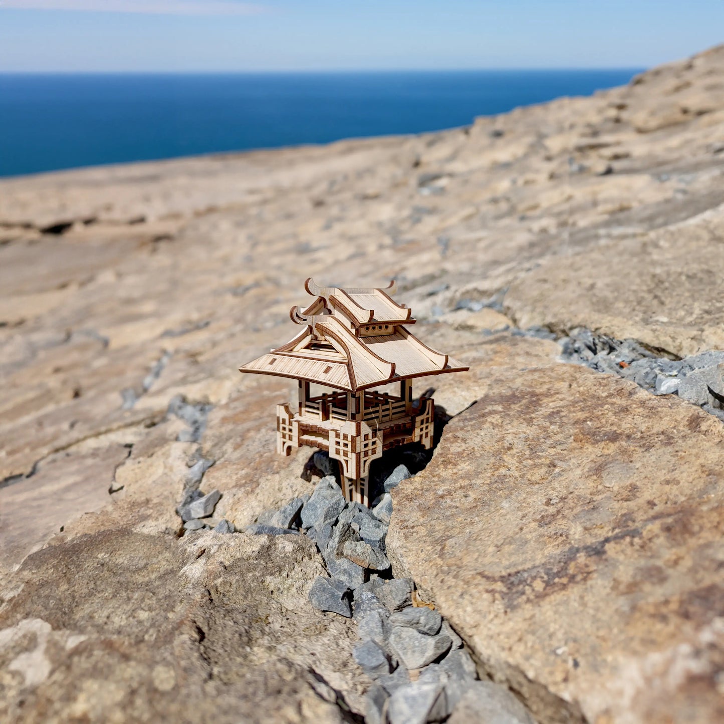 Distant high-angle shot of a laser cut plywood dynamic pavilion model sitting on wide rocky coast with sea view.