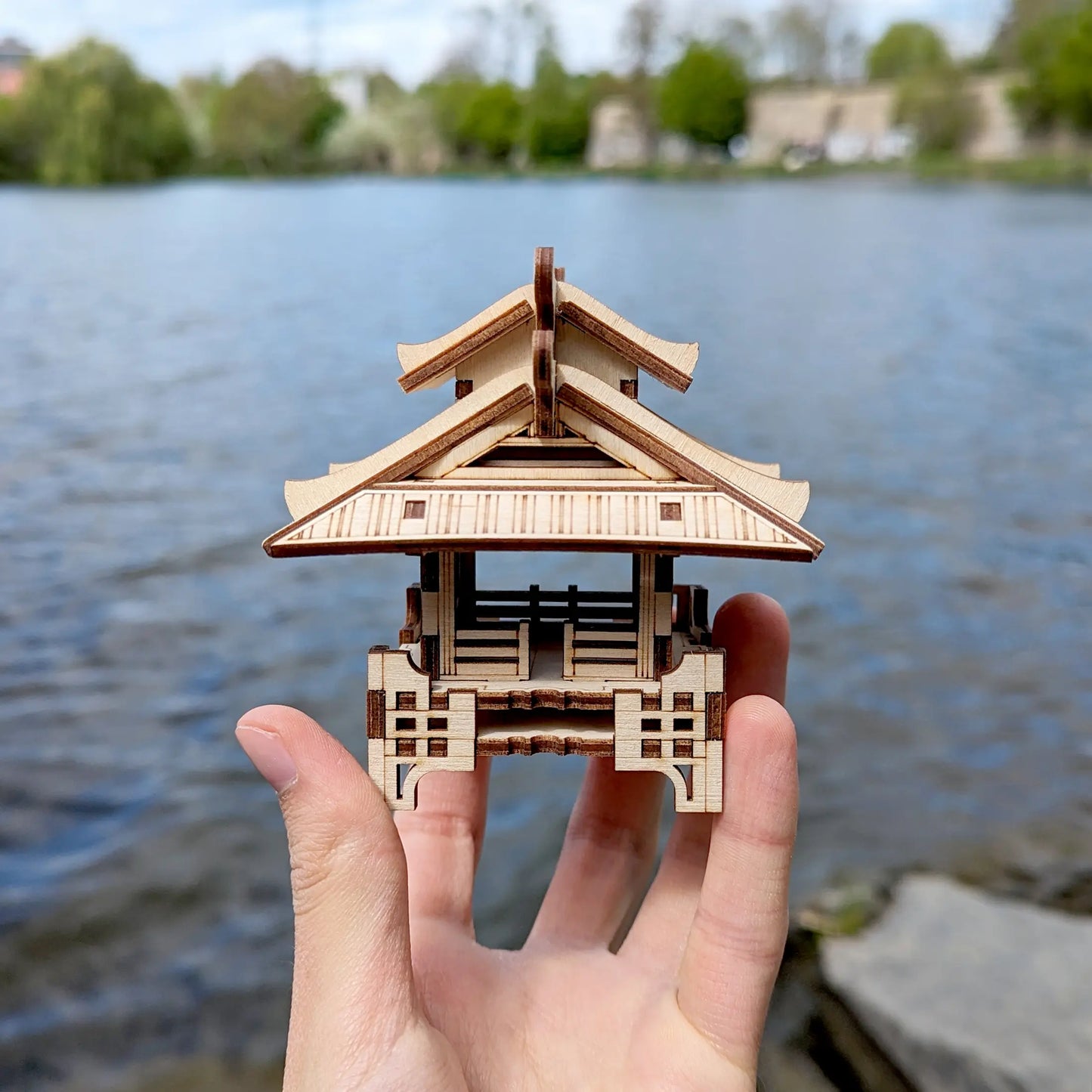 Symmetrical front view of a miniature laser cut plywood shinto shrine held by fingers with a soft blue water backdrop.