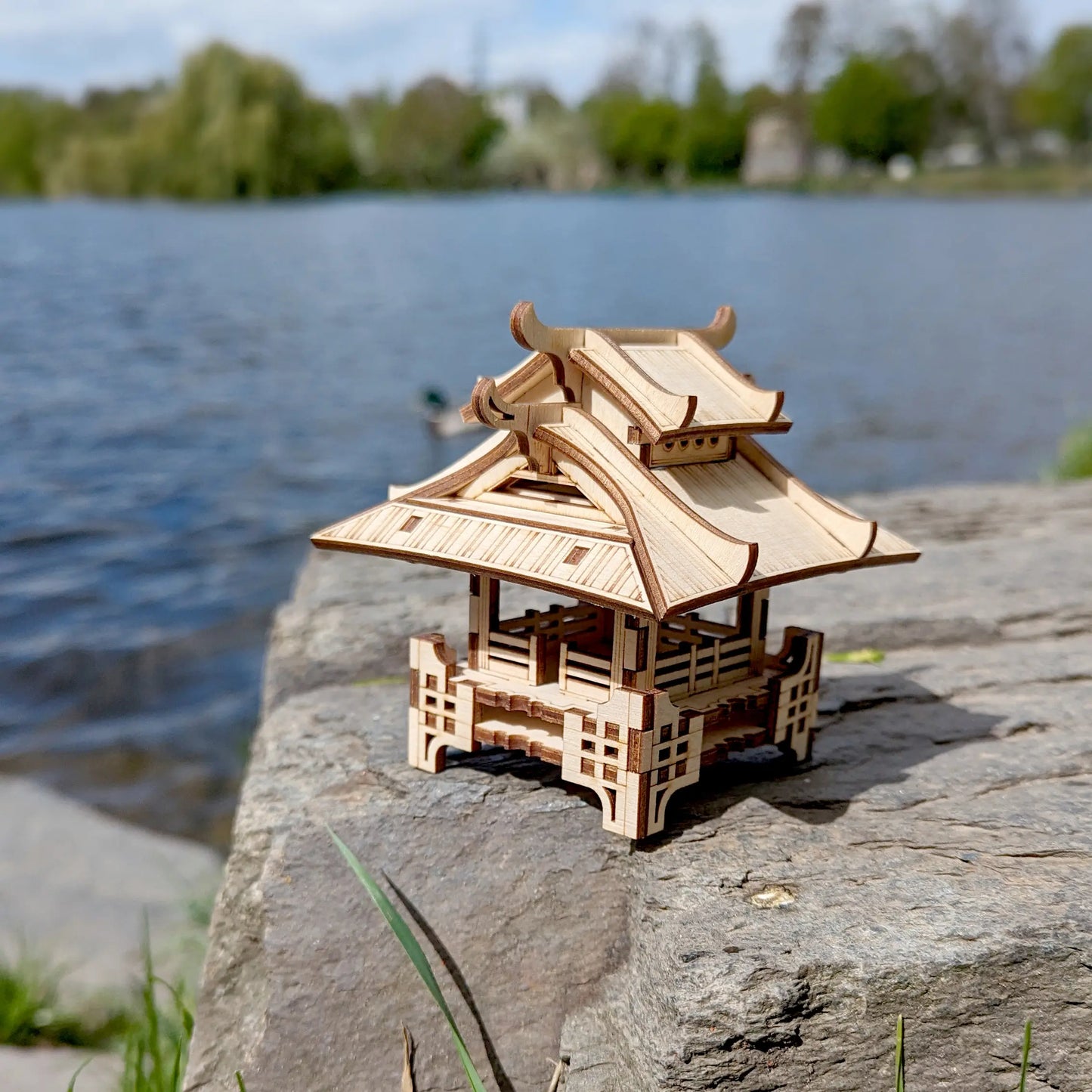 Three-quarter view of a mini laser cut plywood traditional shrine placed on a massive stone next to a flowing river.
