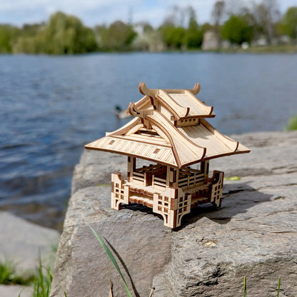 Three-quarter view of a mini laser cut plywood traditional shrine placed on a massive stone next to a flowing river.