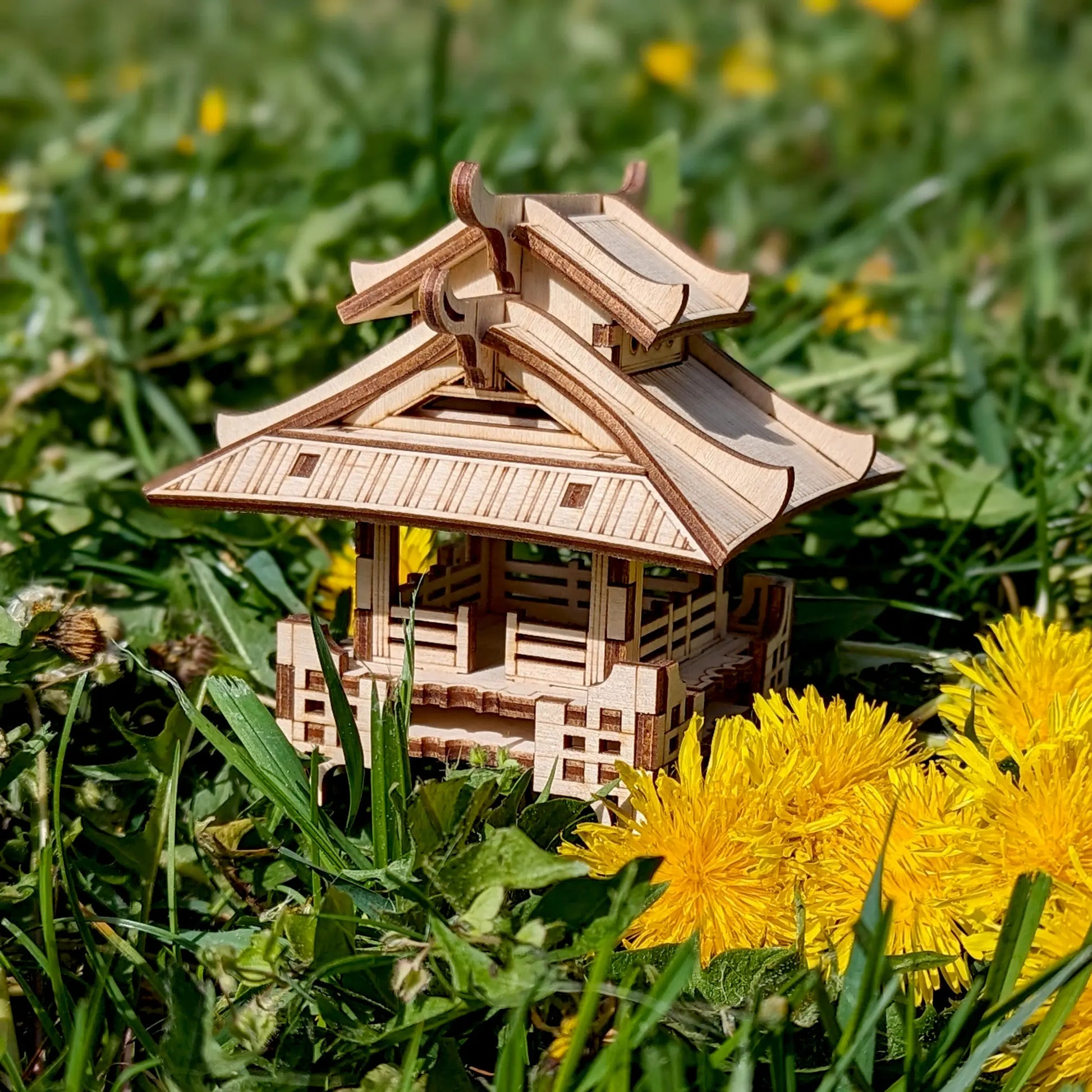 Small laser cut plywood asian house model surrounded by vibrant yellow dandelion flowers and lush green spring grass.