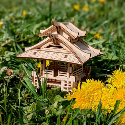 Small laser cut plywood asian house model surrounded by vibrant yellow dandelion flowers and lush green spring grass.