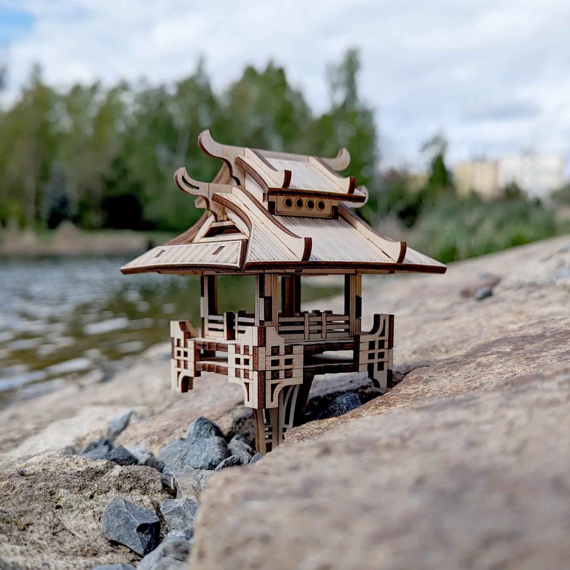 Straight eye-level view of a laser cut plywood wooden pagoda puzzle nestled among grey gravel on a scenic river bank.