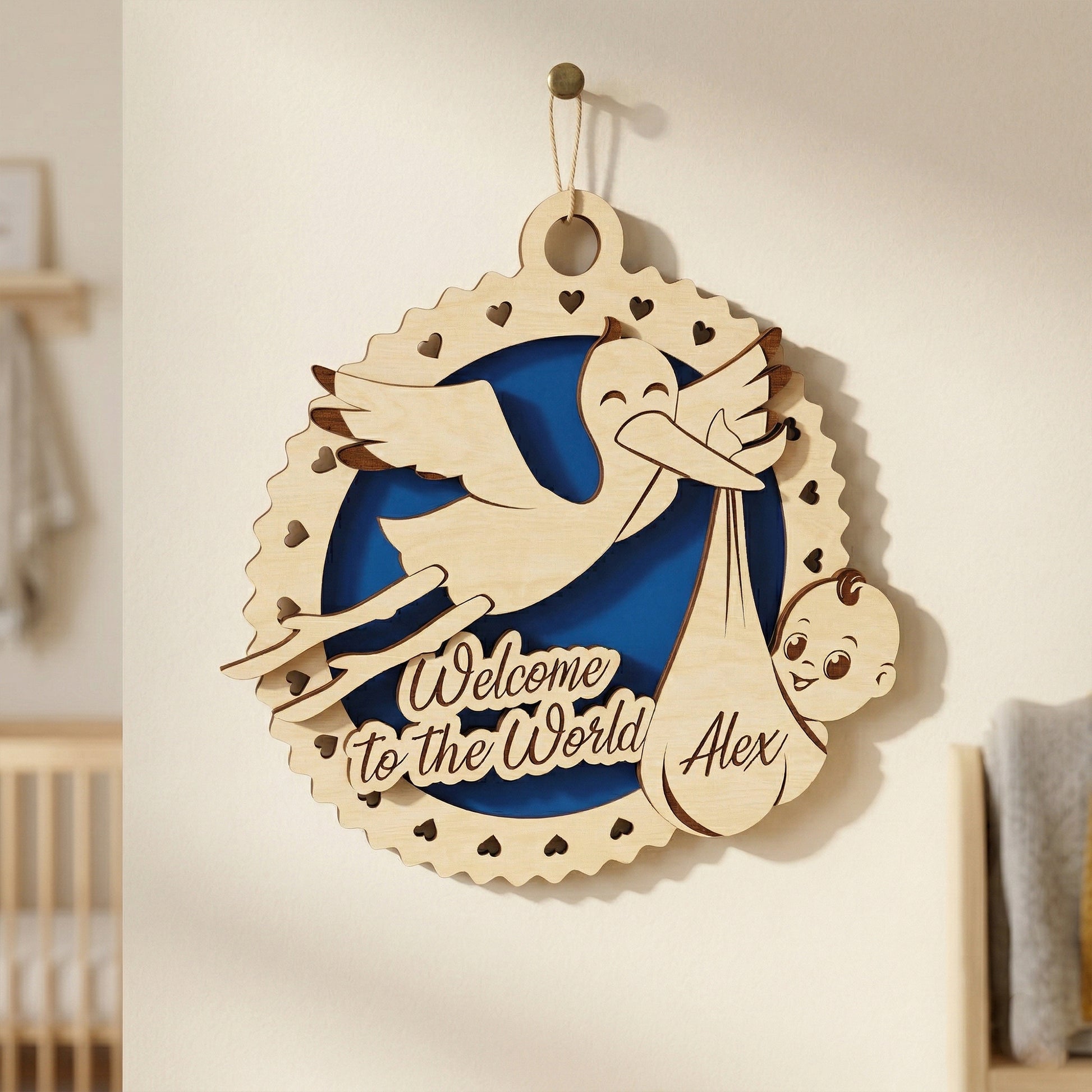 A 3D laser-cut plywood baby birth announcement sign hanging on a nursery wall. The design features a stork carrying a bundle with the name "Alex" and the engraved phrase "Welcome to the World." It includes a blue circular inlay and a scalloped border with heart-shaped cutouts.