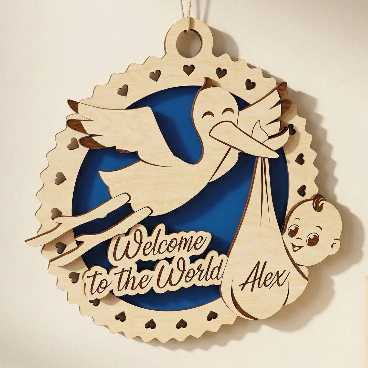 A 3D laser-cut plywood baby birth announcement sign hanging on a nursery wall. The design features a stork carrying a bundle with the name "Alex" and the engraved phrase "Welcome to the World." It includes a blue circular inlay and a scalloped border with heart-shaped cutouts.