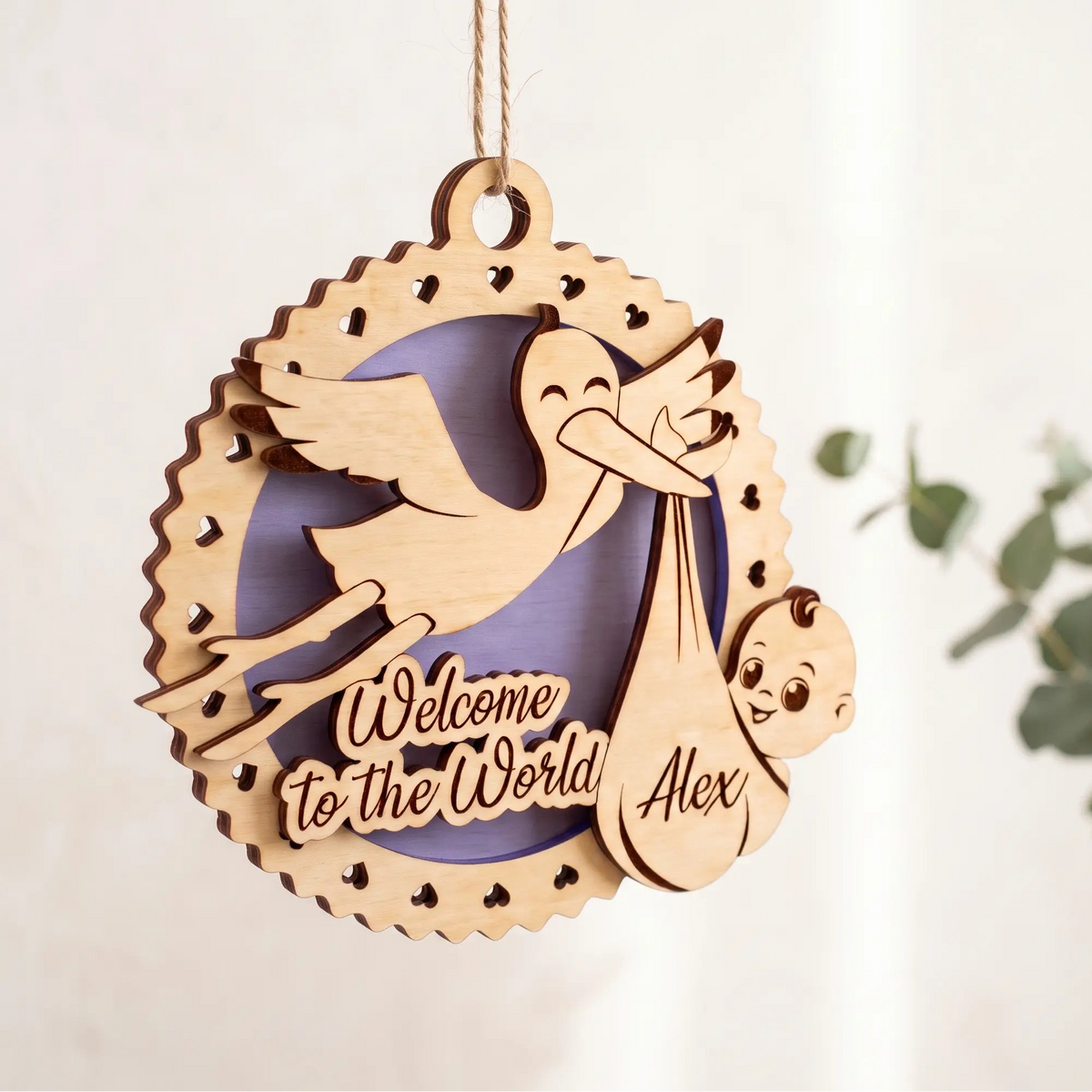 Laser cut plywood newborn baby birth ornament hanging by twine string next to green eucalyptus leaves on light background.