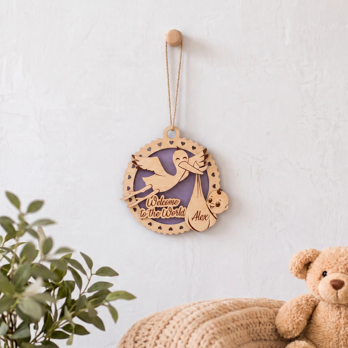 Laser cut plywood baby birth announcement ornament hanging on white nursery wall above knit blanket and plush teddy bear.