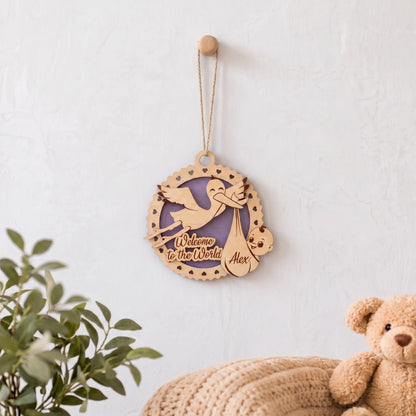 Laser cut plywood baby birth announcement ornament hanging on white nursery wall above knit blanket and plush teddy bear.
