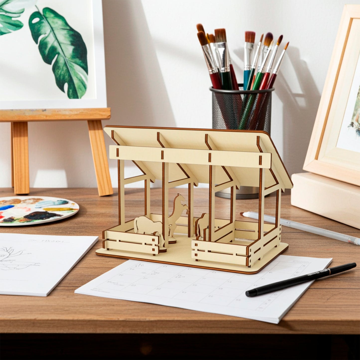 Laser cut plywood wooden barn or stable model featuring a slanted roof and two stalls with silhouette cutouts of horses. The assembled model rests on a desk next to art supplies, brushes, and a window with daylight streaming in.