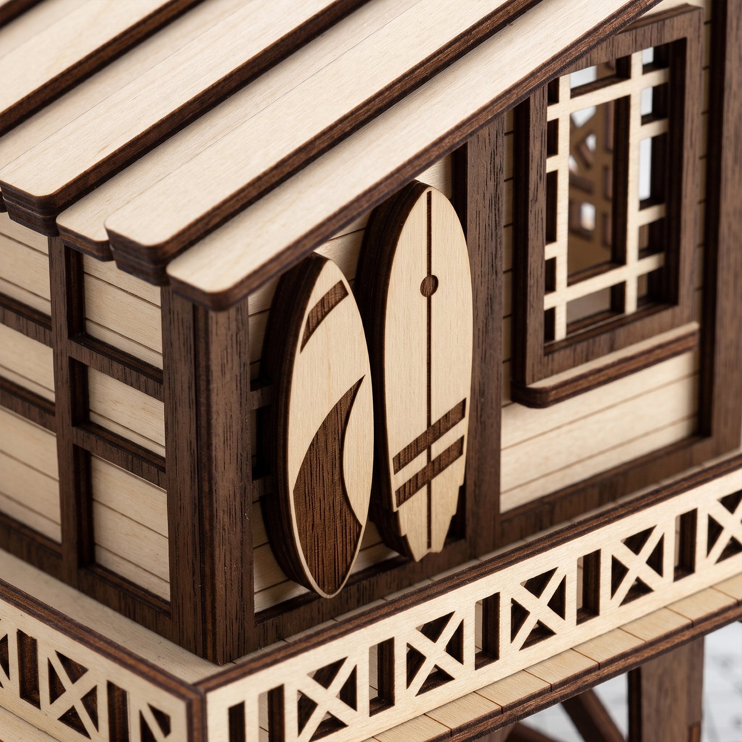 Close-up of a laser-cut birch plywood beach house wall with two miniature surfboards, windows, and dark burnt edges
