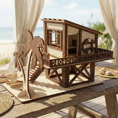 A laser cut plywood beach house model on a rustic deck. It features a stilt base, balcony, palm trees, and lounge chair. In the background, a sunny beach is visible through white curtains.