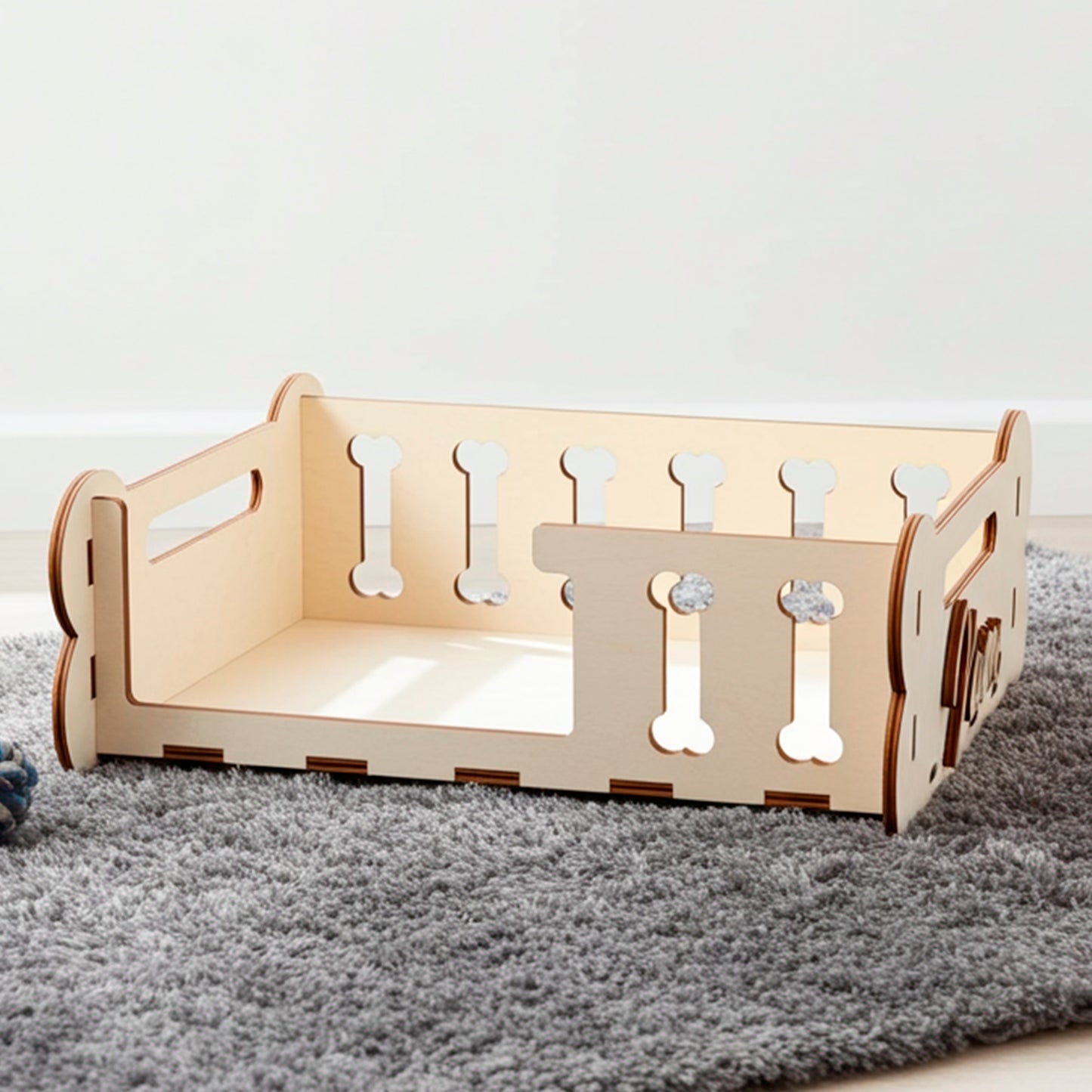 Laser cut plywood wooden Dog Bed frame or storage box with decorative side panels featuring bone-shaped cutouts and personalized engraved text ("Maxie"). The light wood bed, designed as a DIY kit, is displayed on a textured gray rug next to a toy, showing its capacity and function as a pet accessory in a modern interior setting.