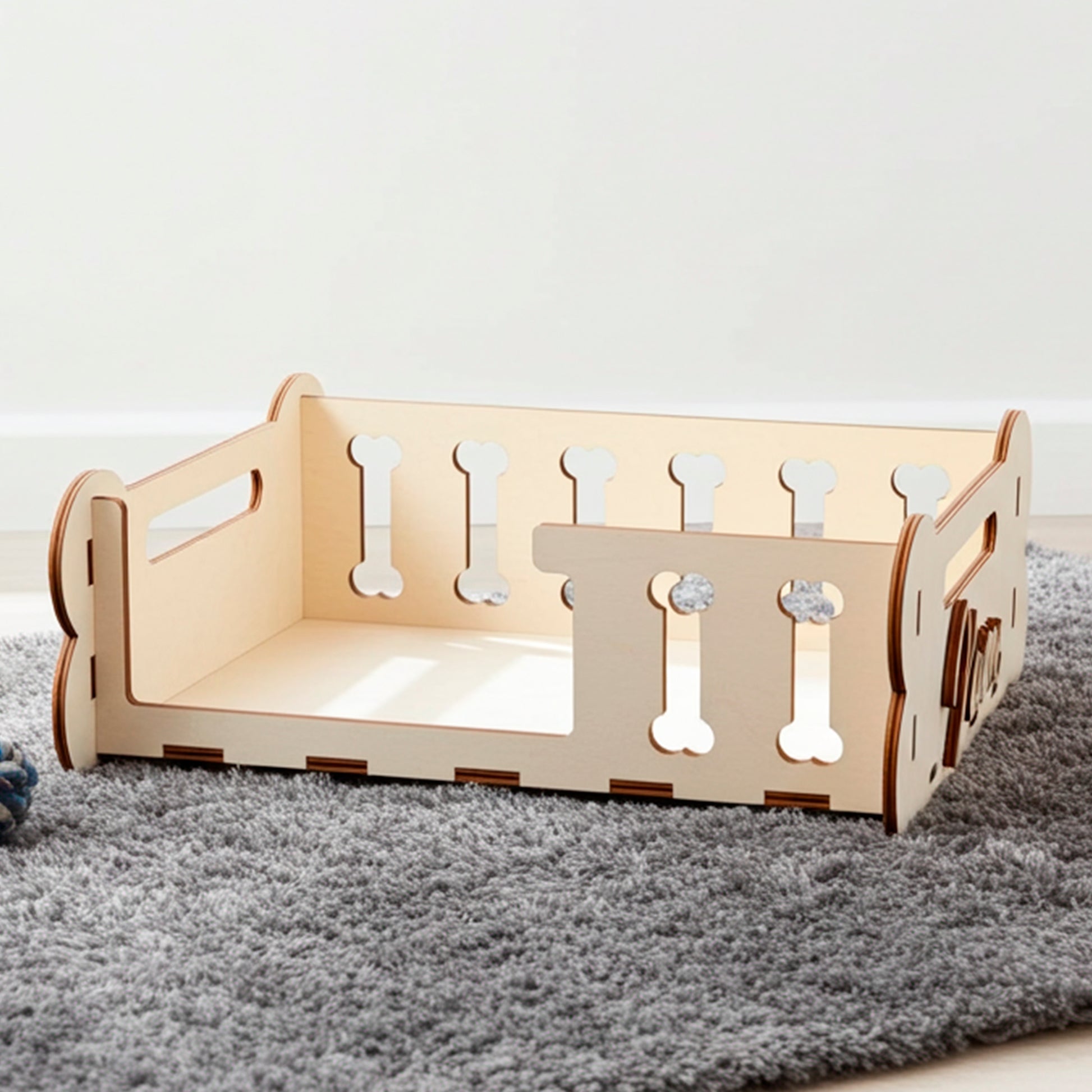 Laser cut plywood wooden Dog Bed frame or storage box with decorative side panels featuring bone-shaped cutouts and personalized engraved text ("Maxie"). The light wood bed, designed as a DIY kit, is displayed on a textured gray rug next to a toy, showing its capacity and function as a pet accessory in a modern interior setting.
