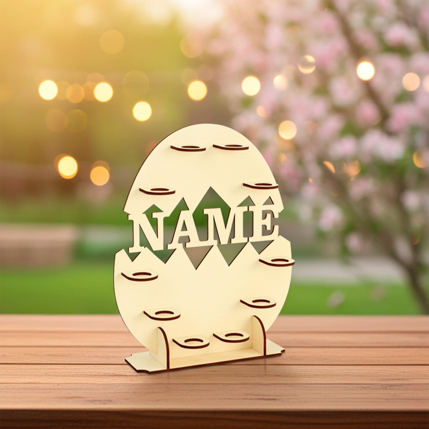 Laser cut plywood wooden Easter egg display stand shaped like a cracked egg, featuring the cutout word "NAME" and many small platforms for holding eggs. The stand sits on a wooden table outdoors with soft bokeh lighting and a pink blooming tree in the background