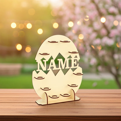Laser cut plywood wooden Easter egg display stand shaped like a cracked egg, featuring the cutout word "NAME" and many small platforms for holding eggs. The stand sits on a wooden table outdoors with soft bokeh lighting and a pink blooming tree in the background
