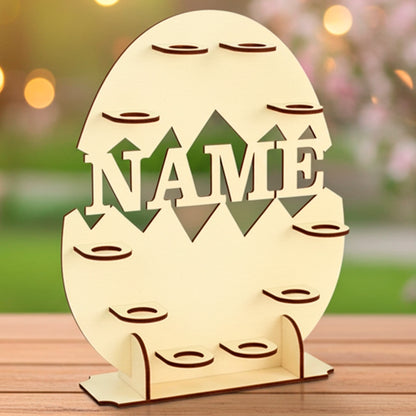 Laser cut plywood wooden Easter egg display stand shaped like a cracked egg, featuring the cutout word "NAME" and many small platforms for holding eggs. The stand sits on a wooden table outdoors with soft bokeh lighting and a pink blooming tree in the background
