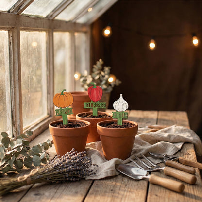 Collection of vibrant laser cut wooden Big Garden Stakes Set in shapes of pumpkin, red bell pepper, and garlic head with engraved names, placed in terracotta pots on a rustic indoor gardening setup background.