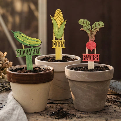 
Set of wooden laser cut Big Garden Stakes featuring colorful designs for cucumber, corn, and radish with bold engraved labels, placed in soil-filled pots against a warm indoor window background.