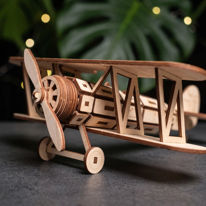 A wooden laser cut model of a biplane, with a propeller and wings, is displayed on a surface with blurred green leaves in the background.