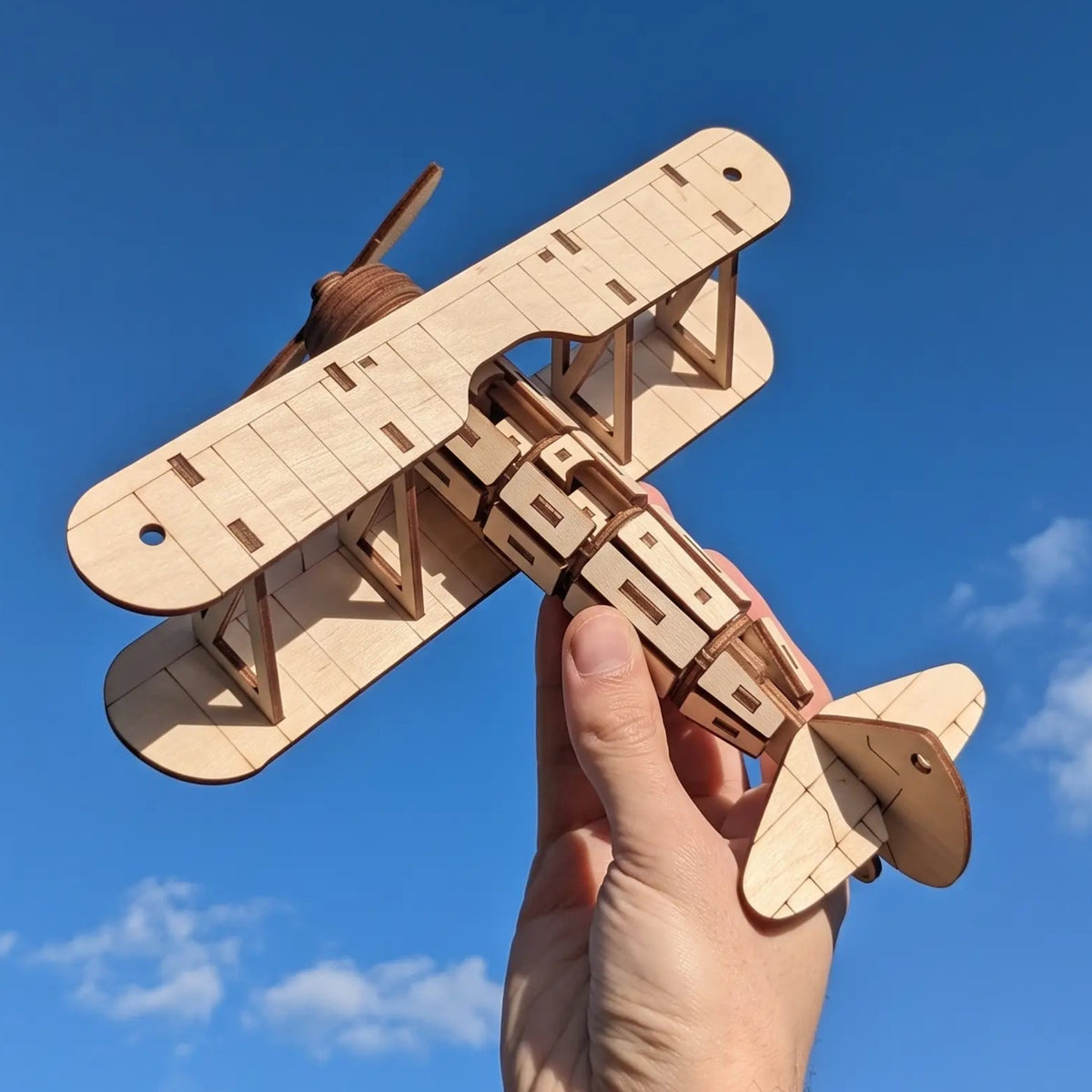 A hand is holding a wooden laser cut model of a biplane against a blue sky with clouds.