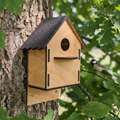 A wooden laser cut birdhouse with a dark gray roof and trim, mounted on a tree trunk outdoors. 