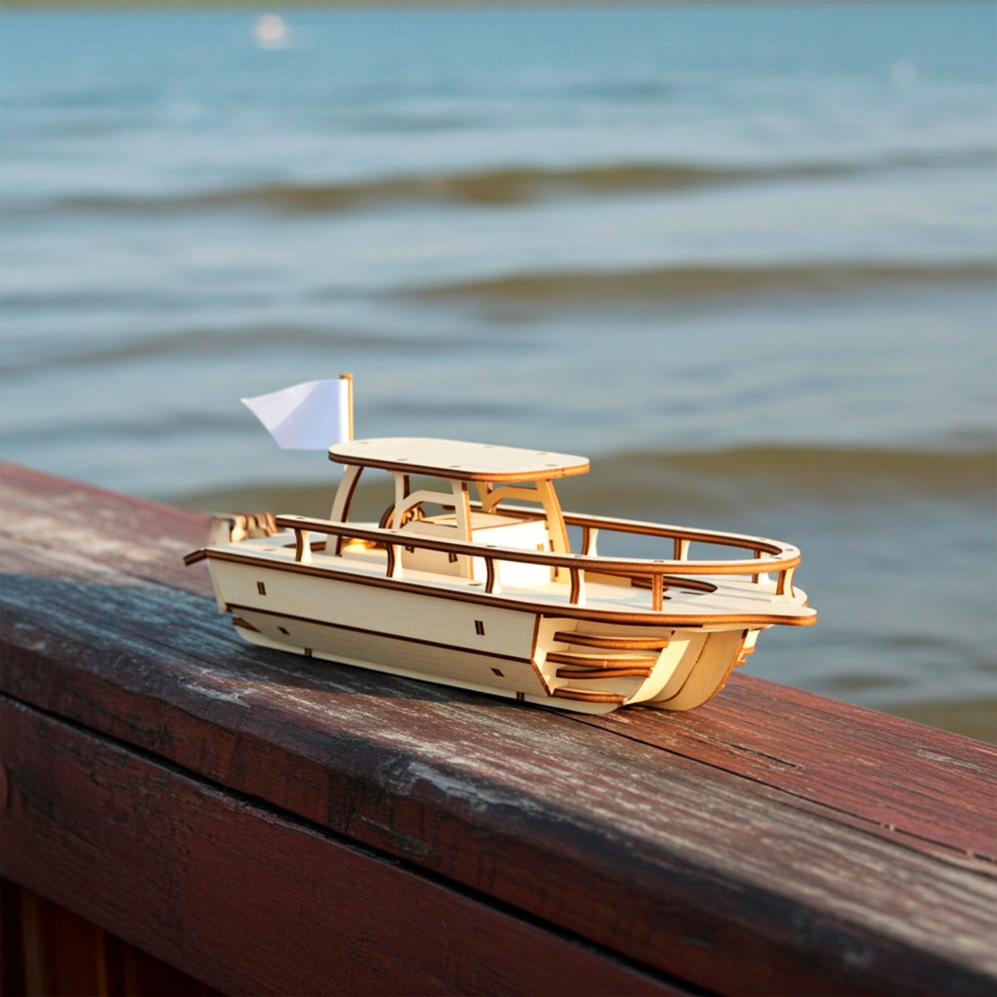 A light wooden  plywood laser cut model of a speedboat with a small white flag, resting on a dark wooden railing, with a blurry blue lake and gentle waves visible in the background.