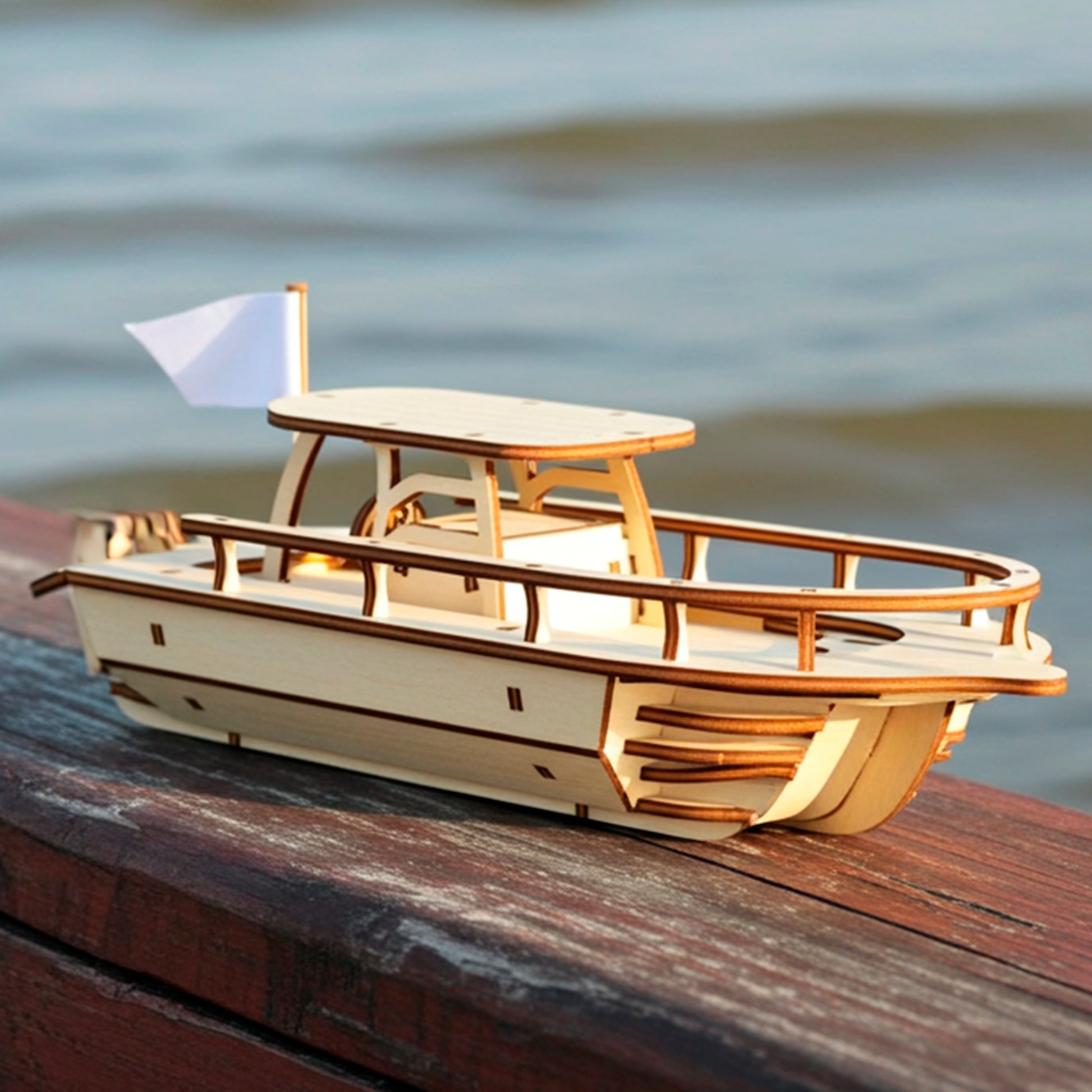 A light wooden  plywood laser cut model of a speedboat with a small white flag, resting on a dark wooden railing, with a blurry blue lake and gentle waves visible in the background.