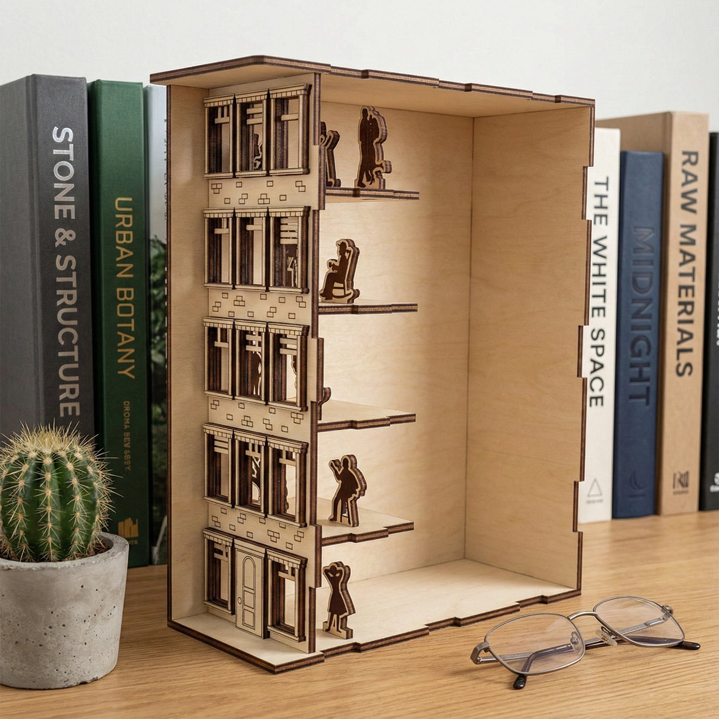 Side view of a laser cut wooden Book Nook Apartment House showing internal floors and silhouettes, placed on a wooden desk with a minimalist study background.