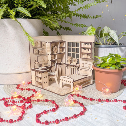 Laser cut wooden miniature bedroom scene displayed among potted plants. The tiny desk, chair, bookshelves, bed, and window details are softly lit with string lights and surrounded by red bead garland