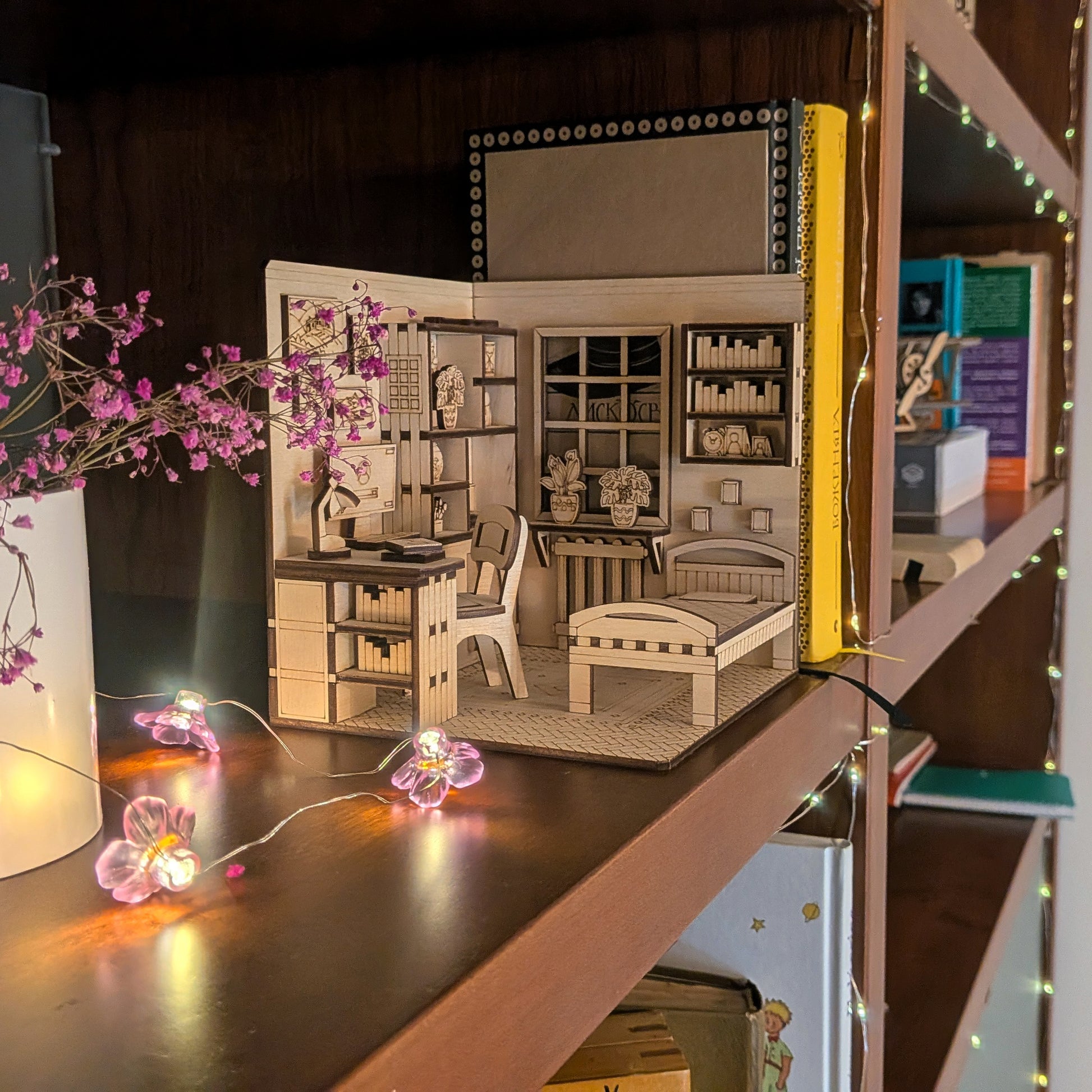 Miniature model of a laser cut book nook room with furniture and decor, illuminated by string lights.