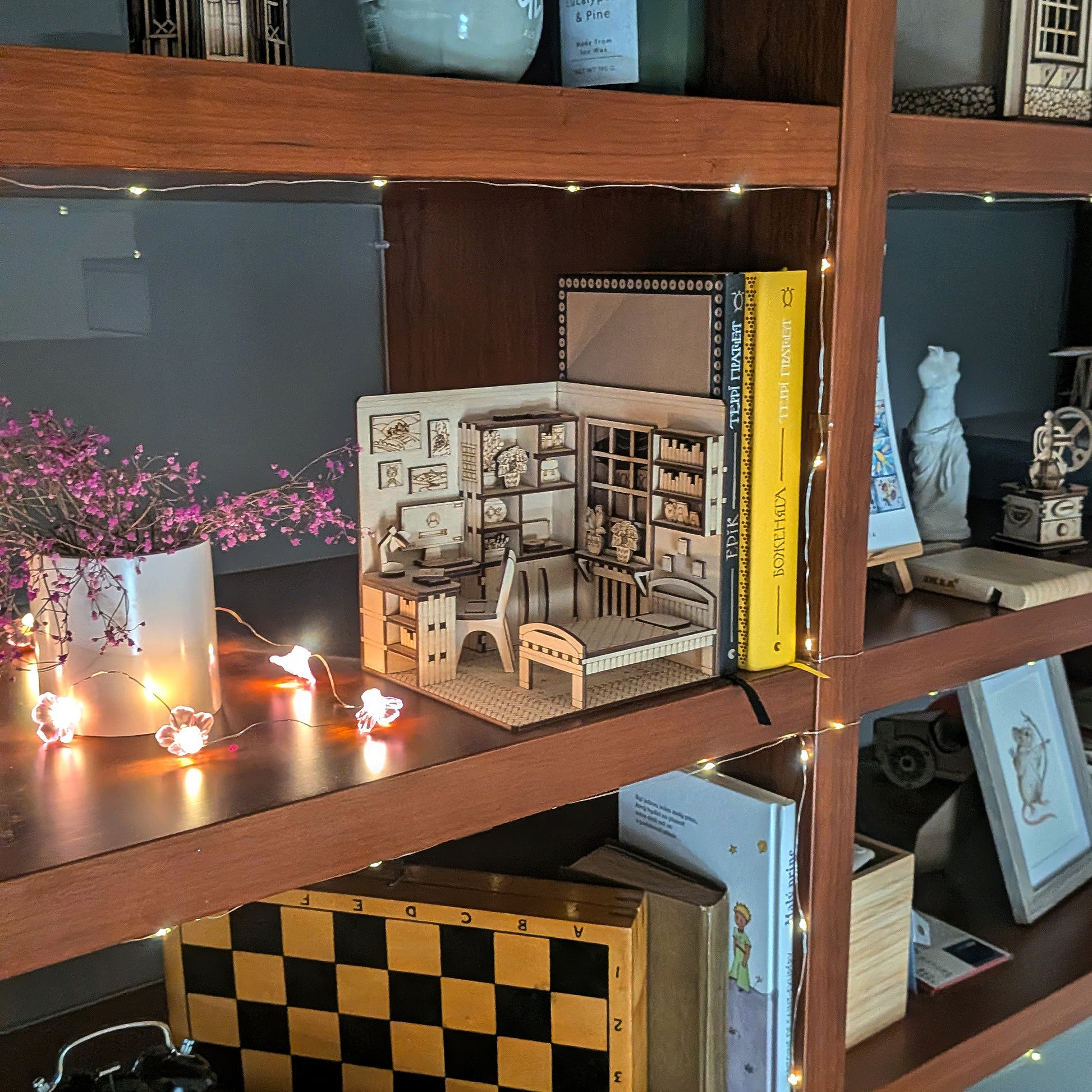 Laser cut wooden miniature bedroom book nook displayed on a bookshelf. The scene includes a small desk with a chair, bookshelves, plants, and a bed, softly lit with warm fairy lights beside a vase with dried flowers